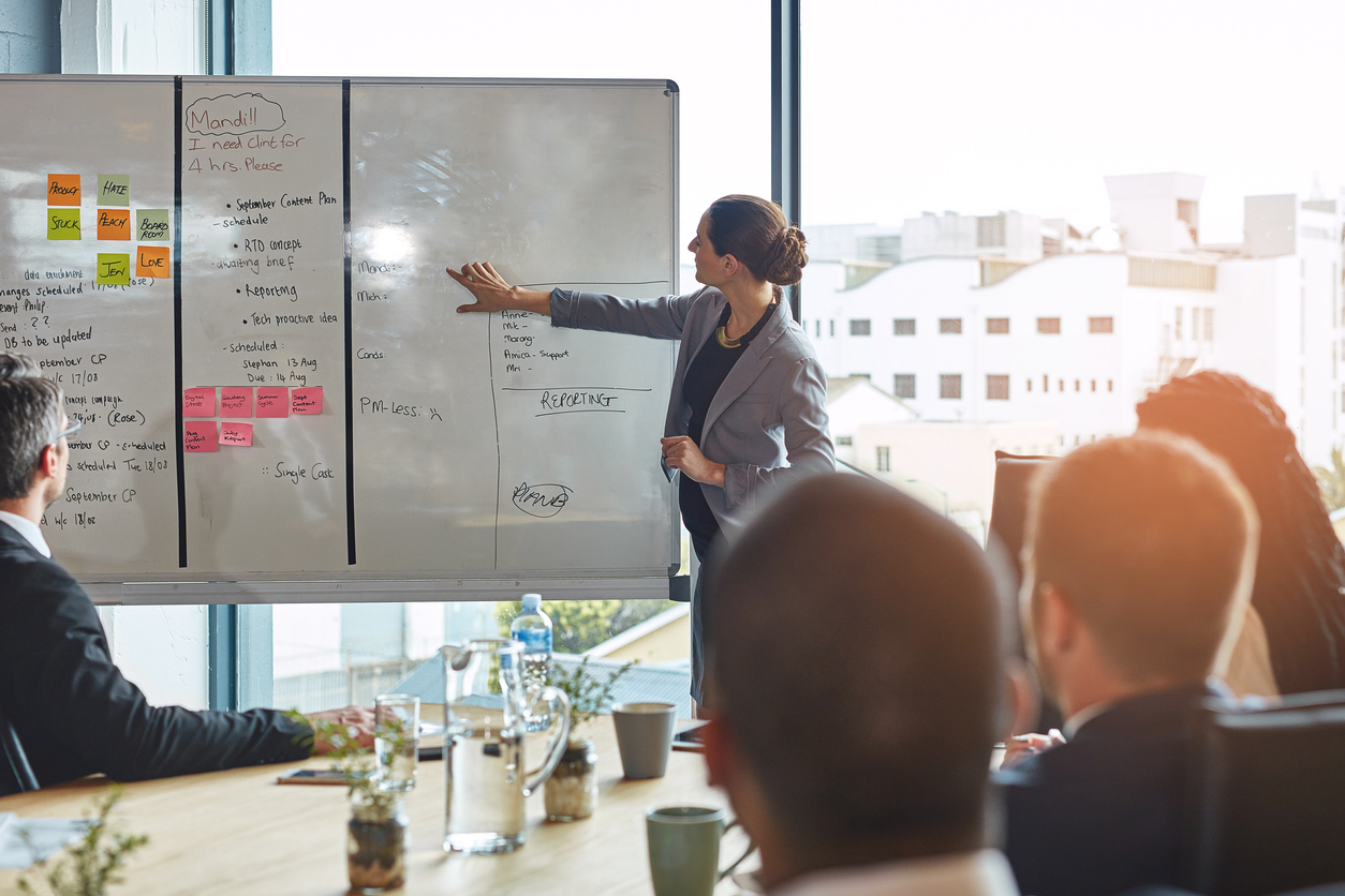 Leader presenting a sticky-note plan on a whiteboard to a seated team.